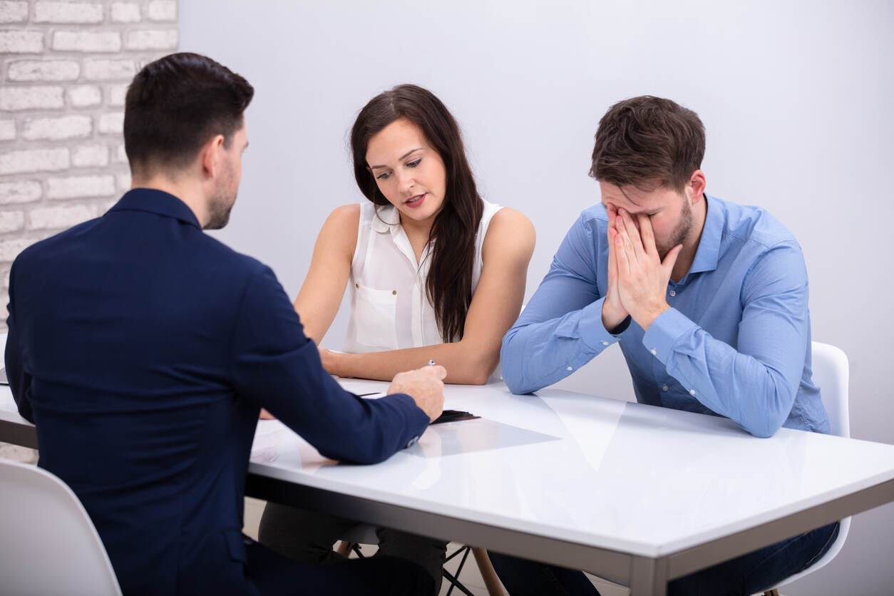 A distressed man covers his face at a table with a mediator and a woman, illustrating how divorce mediation can help couples avoid the emotional drama of court.