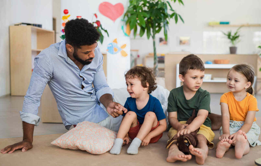 A father sitting with four young children, two of whom are crying, while the other two look on. The father appears to be engaged in a conversation with one of the children, offering guidance or comfort.