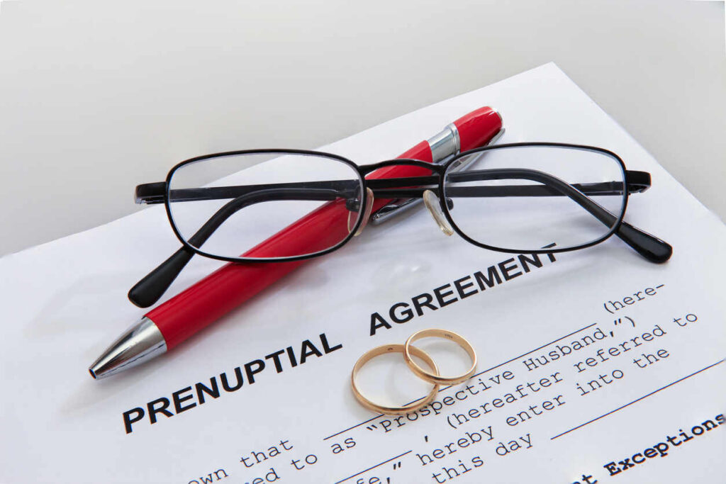 Close-up of a prenuptial agreement document with a red pen, eyeglasses, and two gold wedding rings.